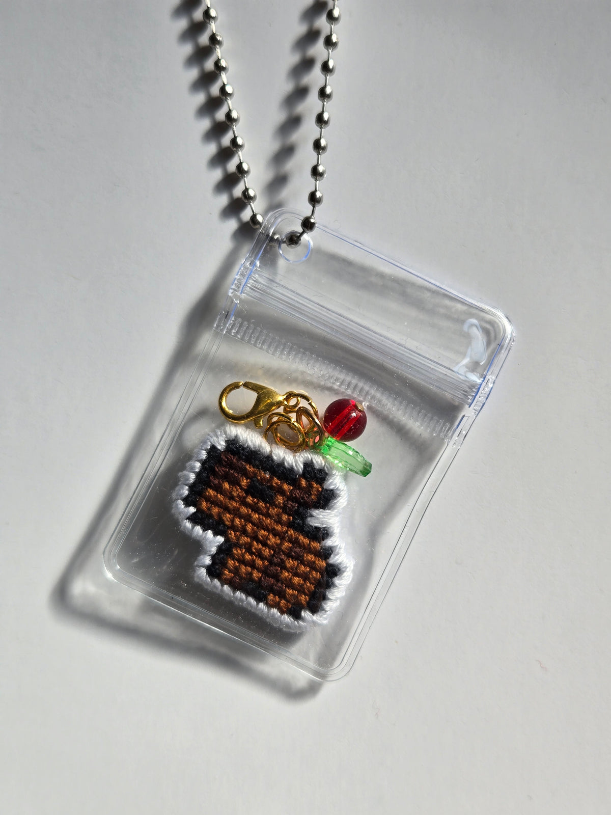 A small clear zip pouch pendant necklace on a silver ball chain, containing a brown and white pixel-art style charm with a gold clasp, a red bead, and a green bead, photographed on a light gray background.