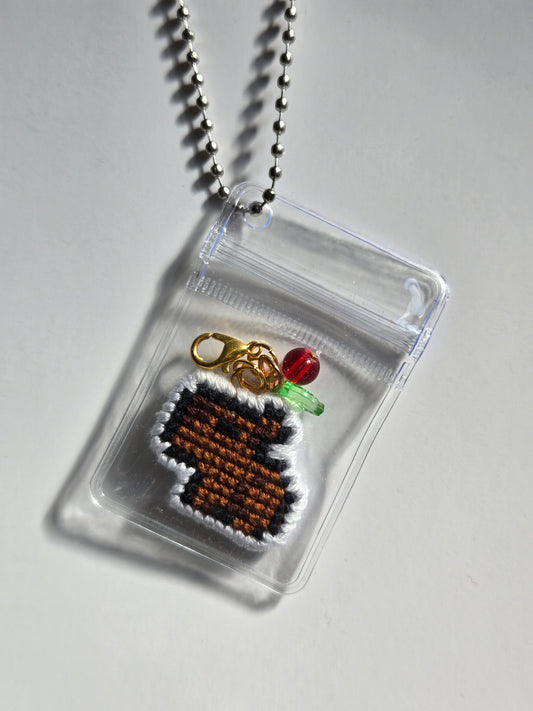 A small clear zip pouch pendant necklace on a silver ball chain, containing a brown and white pixel-art style charm with a gold clasp, a red bead, and a green bead, photographed on a light gray background.